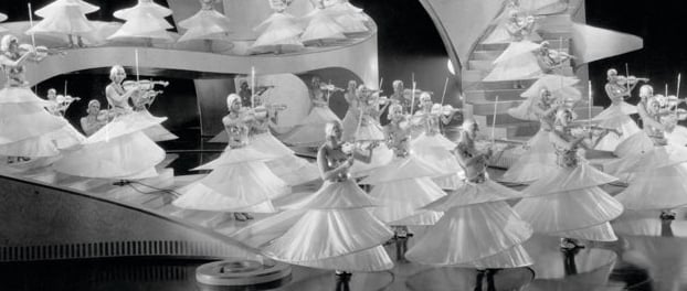 Female dancers in circular costumes playing white violins on a black‑and‑white Busby Berkeley set.