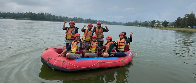 Delapan orang bersemangat di perahu karet merah melakukan rafting di Situ Cileunca Bandung, mengenakan jaket pelampung oranye