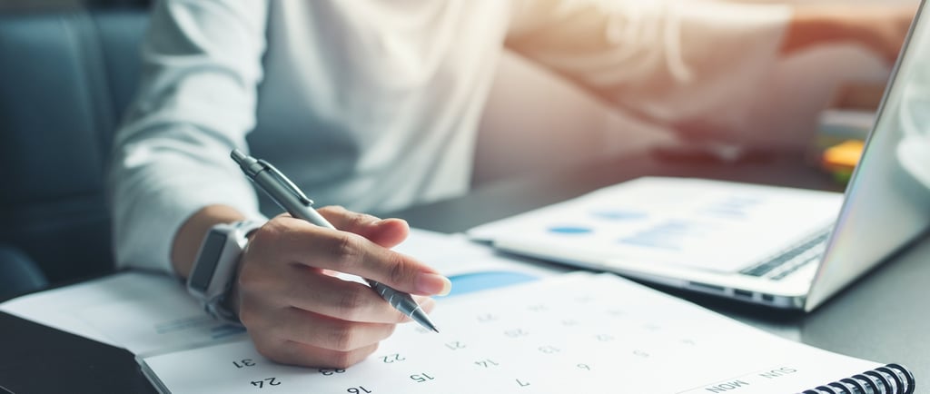someone working at a desk with a calendar, laptop, and pen in hand