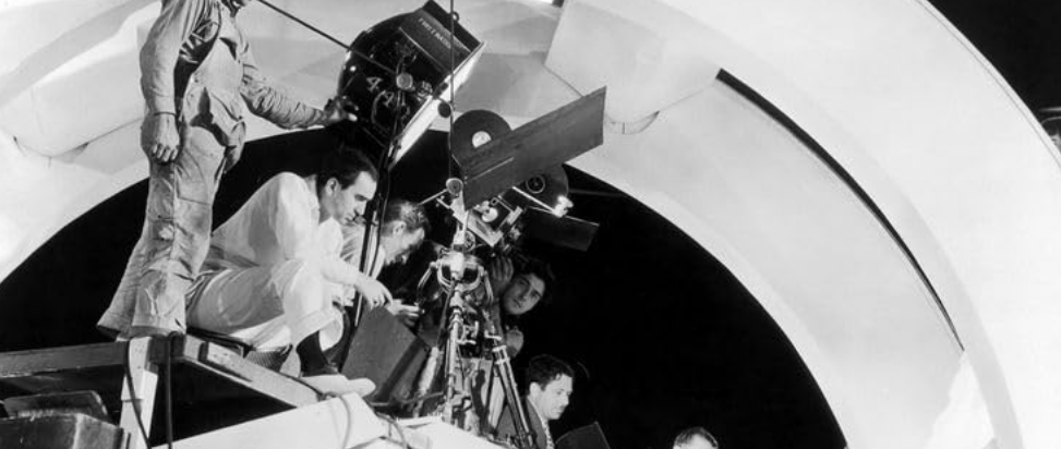 Busby Berkeley on a tall filming platform with cameras and lights, directing dancers below.