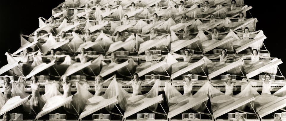 Multiple rows of white grand pianos with white‑clad chorus girls in a Busby Berkeley black‑and‑white