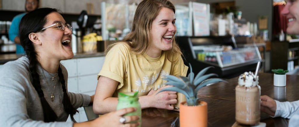 Dos jóvenes sonríen y conversan en una cafetería moderna, disfrutando bebidas y representando la cul