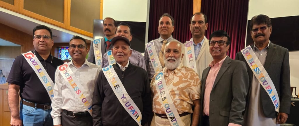 Nine South Asian fathers and father-figures wearing celebratory sashes, standing and smiling under a