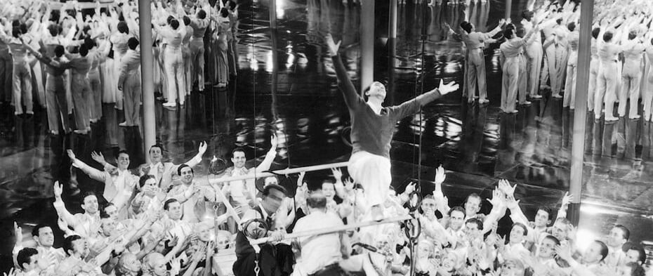 Busby Berkeley demonstrating arm movements to dancers on a mirrored stage platform.