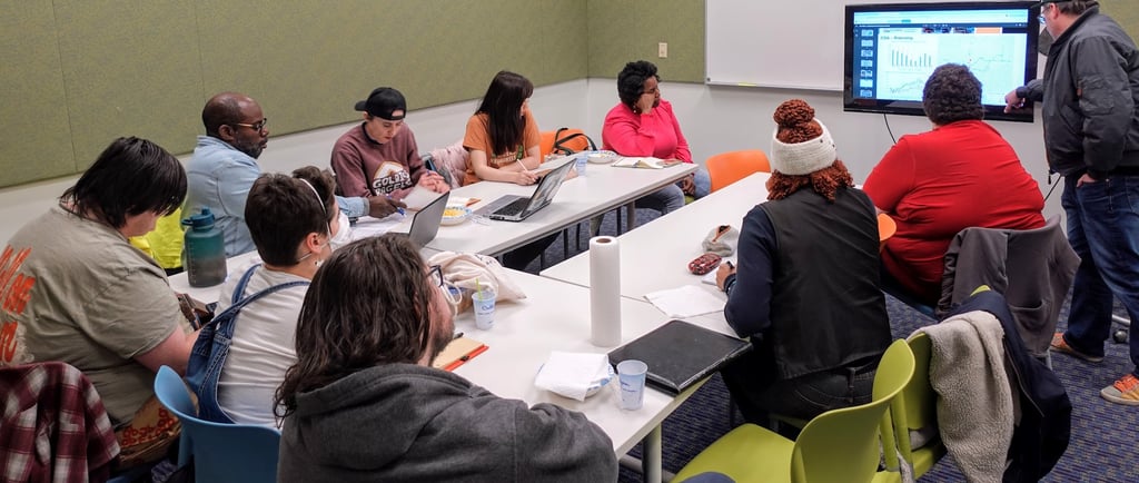 A group of people gathered around a table, looking intently at a presenter in the front of the room