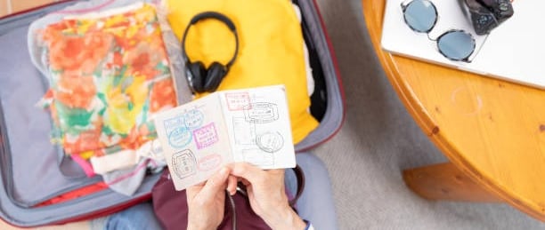Overhead view of a person holding an open passport displaying travel stamps, next to an open suitcas