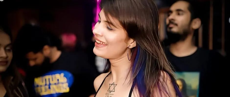 Smiling woman with purple hair highlights and a chest tattoo dancing at a crowded night club party.