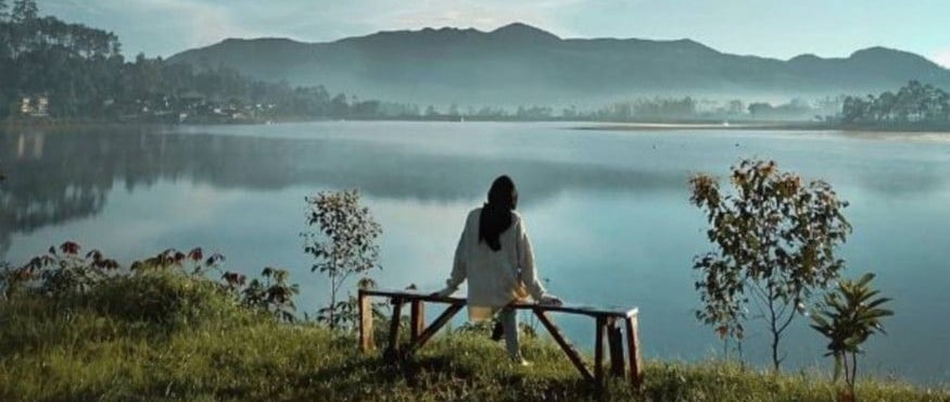 Seorang wanita berhijab duduk di bangku kayu menghadap danau Situ Cileunca yang tenang dengan latar belakang pegunungan dan l
