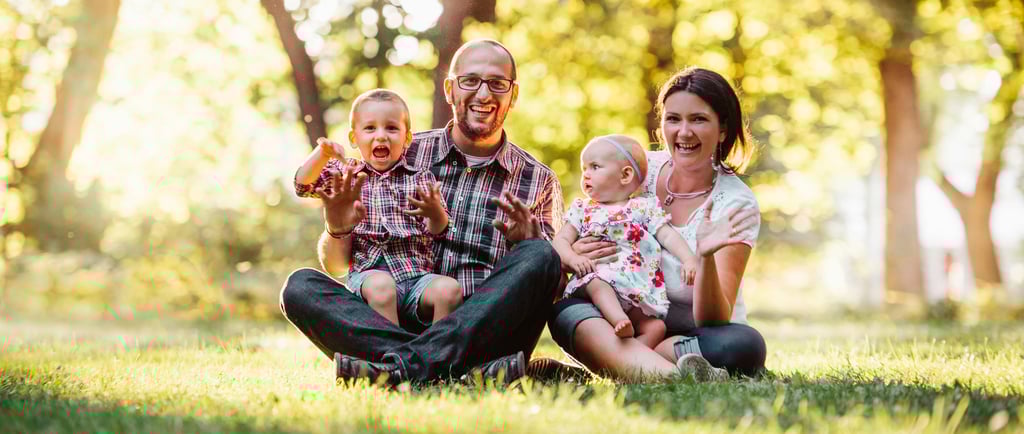 homem e mulher sentados em um gramado, cada um segura uma criança, expressão de felicidade no rosto