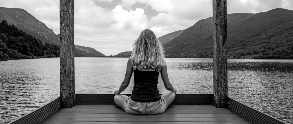A woman practices yoga meditation on a wooden pier overlooking a scenic mountain lake.