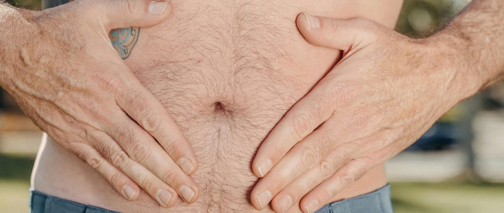 Close‑up of an adult male abdomen with hands resting on the stomach, showing natural body hair and a side tattoo, outdoors