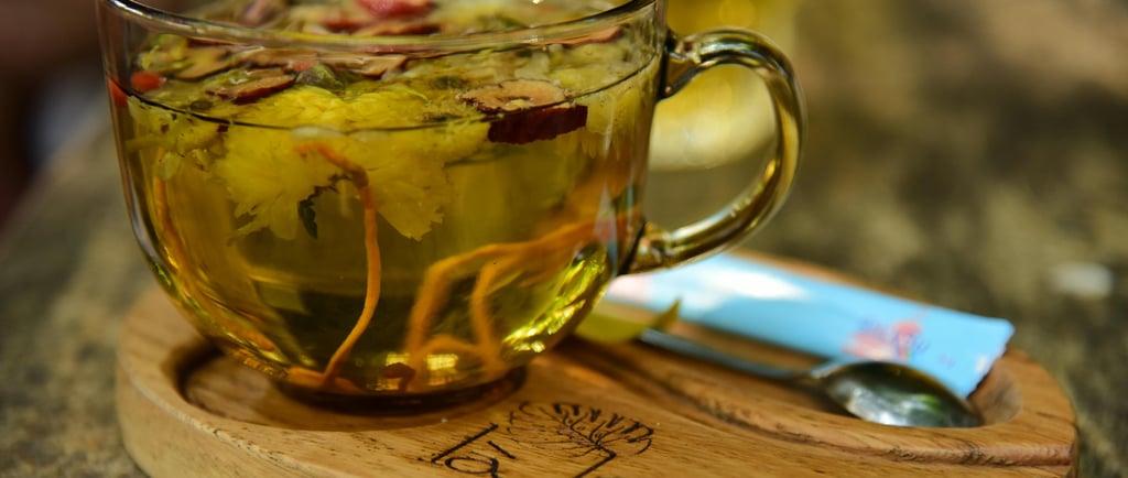 “Glass cup of herbal tea with visible flower blossoms on a wooden saucer, placed beside a spoon and a blue packet 