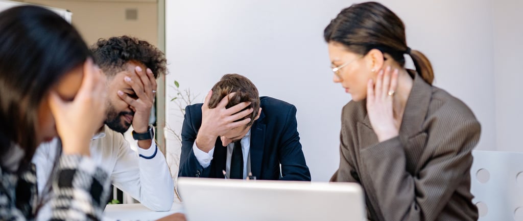 Four people sit around a table in an office meeting, with three of them resting their heads in their hands