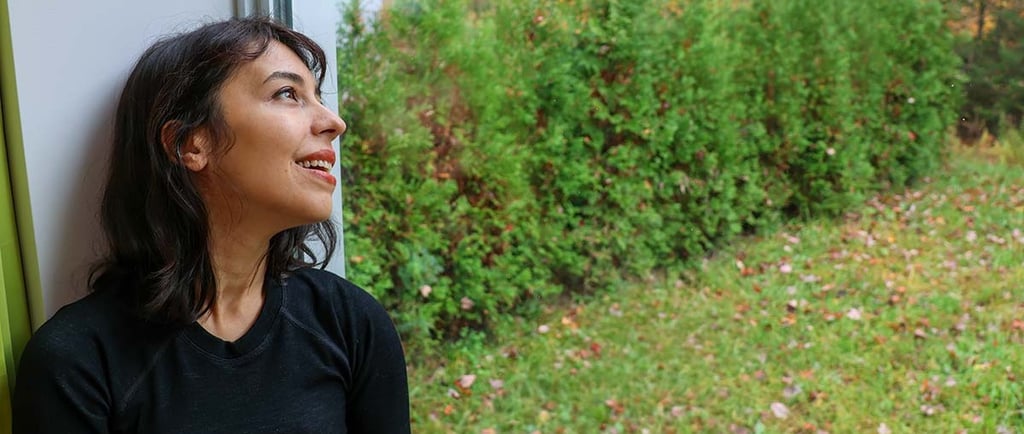 Woman sitting by a window looking outside at evergreen trees and autumn foliage.
