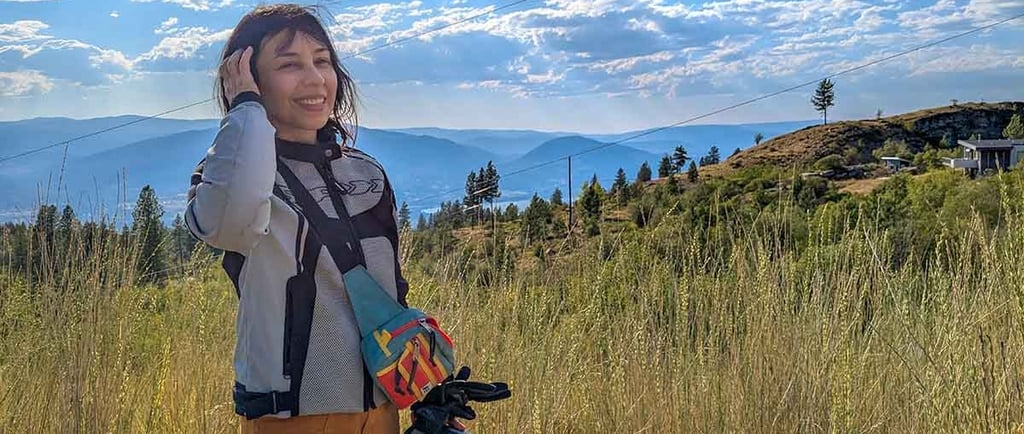 Woman standing beside a motorcycle helmet overlooking mountains and grasslands near Penticton, British Columbia.