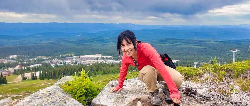 Woman crouching on a rocky mountain viewpoint overlooking Big White and surrounding forests under cloudy skies.