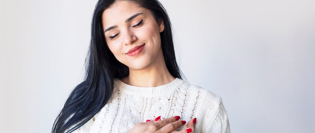 Mulher sorridente com os olhos fechados, vestindo um suéter branco de tricô e com as mãos no coração