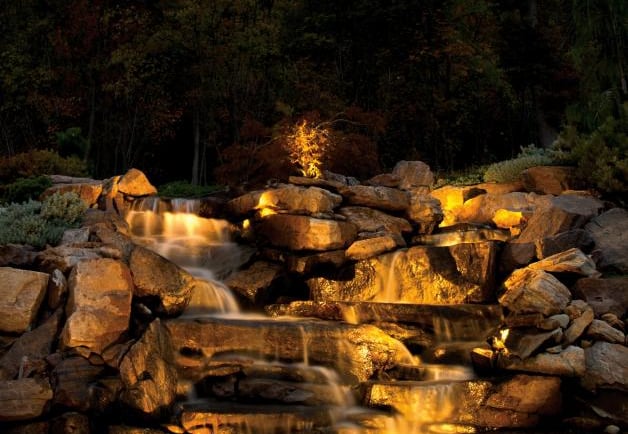 Beautiful, illuminated rock waterfall with lush trees, creating a serene and natural garden landscape at dusk.