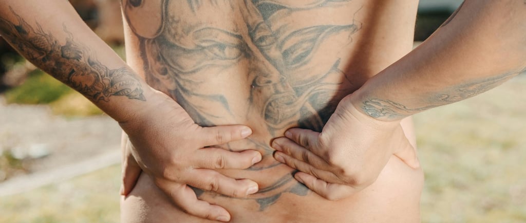 A man with a large back tattoo holds his lower back in pain, signifying chronic lumbar muscle strain.
