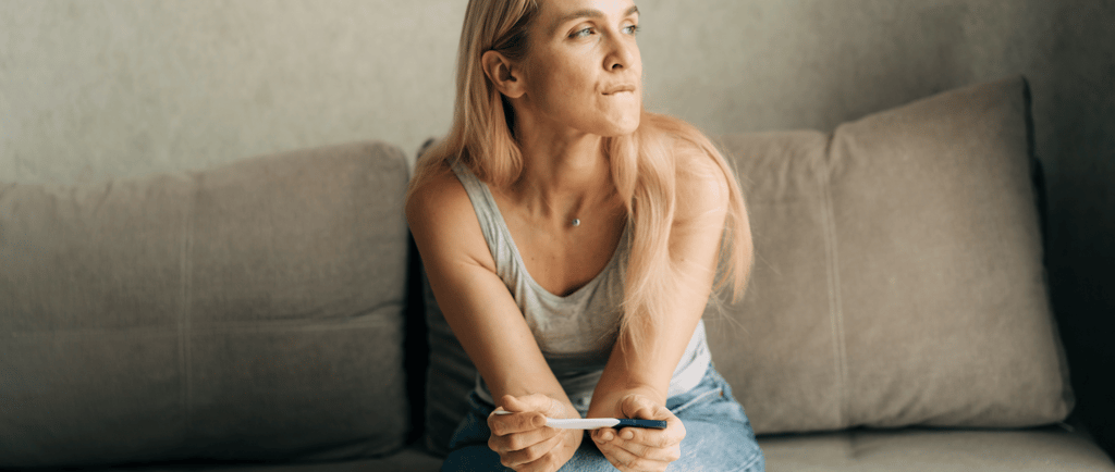 Woman sitting on couch holding a pregnancy test, reflecting on fertility questions