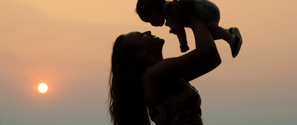 Mother holding child up in the air during sunset, both silhouetted against the sky