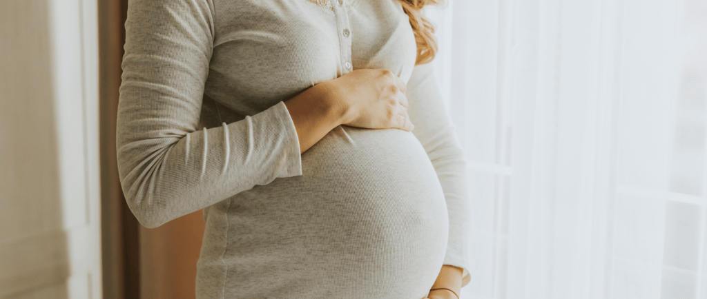 Pregnant woman standing by window with hands on belly