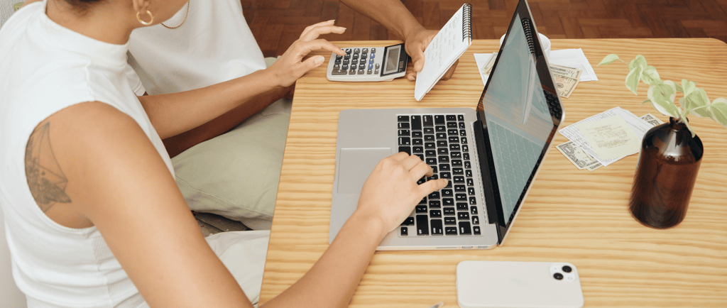 Couple working at a laptop with phone, papers, and calculator on a wooden desk