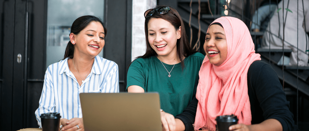 Three women smiling and looking at laptop while enjoying coffee outdoors