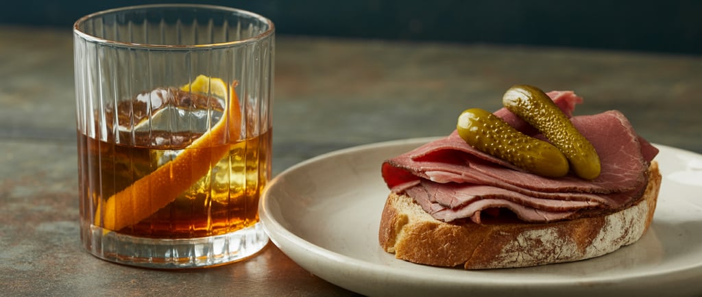 A close-up photograph featuring a classic Rum Old fashioned cocktail and a deli sandwich. 