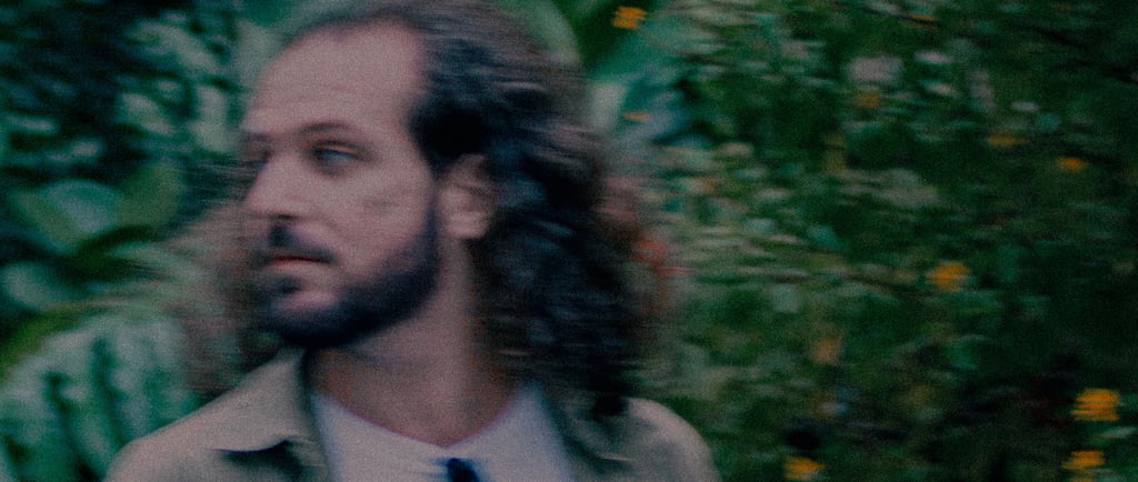 Candid portrait of a bearded man with long hair looking away against a blurred garden background.