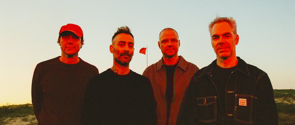 Four members of a rock band standing outdoors at sunset with a red flag in the background.