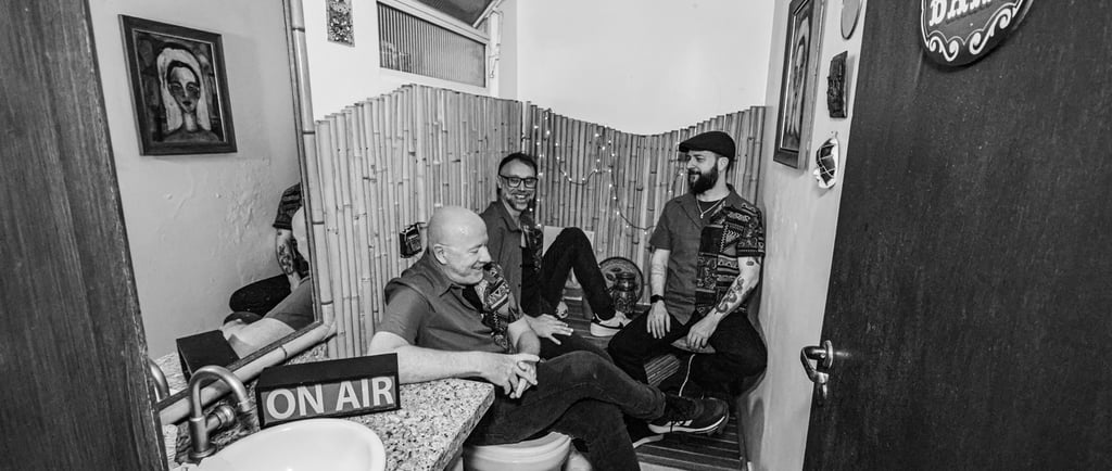 Three men talking in a bathroom-themed studio with an On Air sign and bamboo wall decor.