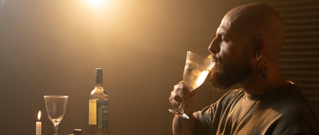 A bearded man drinking white wine from a glass at a candlelit table with a bottle.