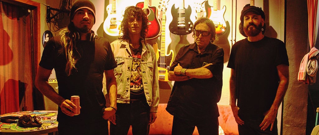 A four-member rock band stands in a music studio with electric guitars mounted on the wall behind them.