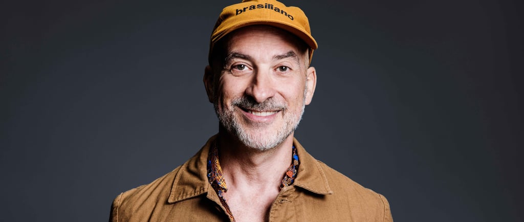 A smiling man with a beard wearing a yellow Brasiliano cap and a tan jacket against a dark background.
