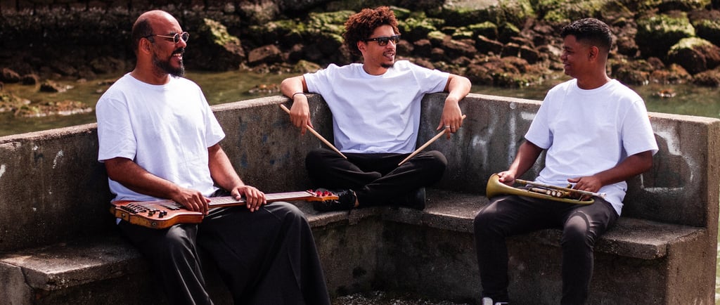 Three musicians holding instruments outdoors sitting on a stone bench near a rocky shoreline.