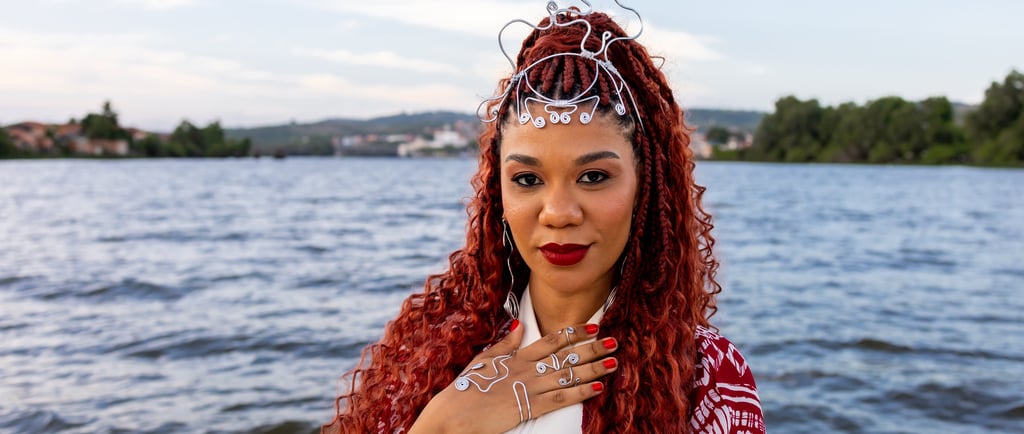 Woman with red braids and silver wire jewelry posing by a scenic river at sunset.