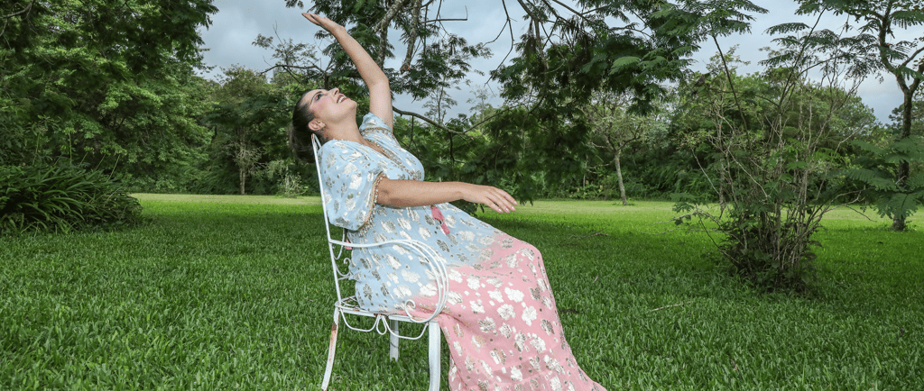 Woman in floral kaftan dress posing on white chair in a lush green garden.