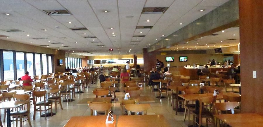 Spacious modern cafeteria with wooden tables and chairs, bright ceiling lighting, and large windows.
