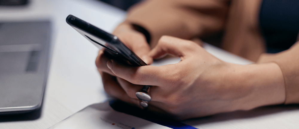 Mulher com um celular nas mãos, um papel e um notebook