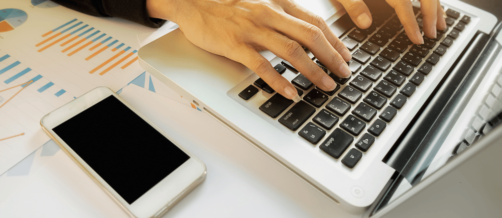 Mulher digitando em um notebook com um celular ao lado