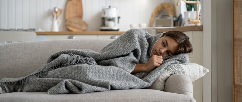 Frau liegt eingehüllt in eine graue Decke auf einem Sofa und ruht sich aus, im Hintergrund eine gemü