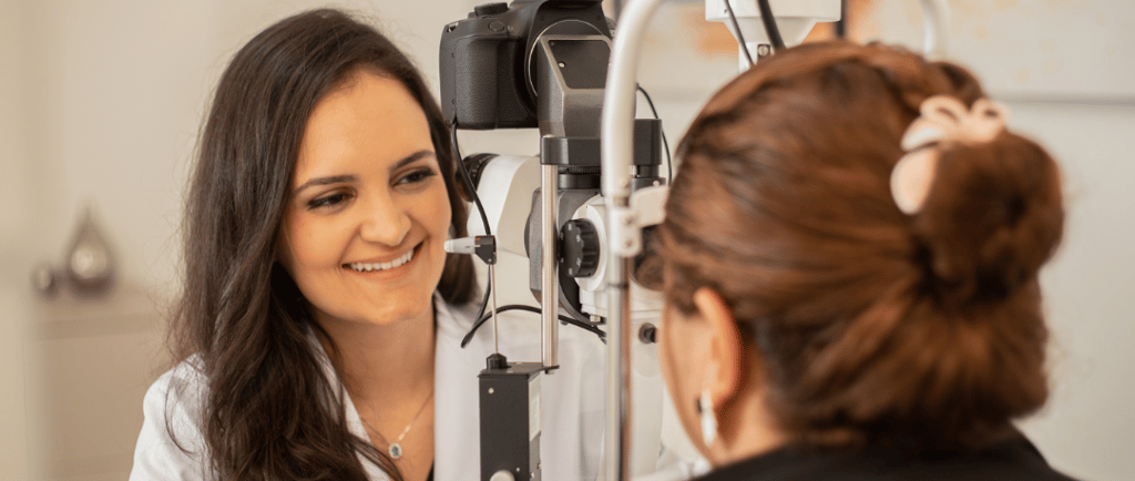 Dra Bruna Melchior de jaleco branco examinando paciente em aparelho de oftalmologia