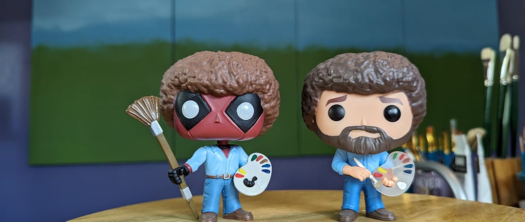 Funko POP Deadpool Bob Ross and Bob Ross figurines standing in front of a 4-panel project painting in an art studio.