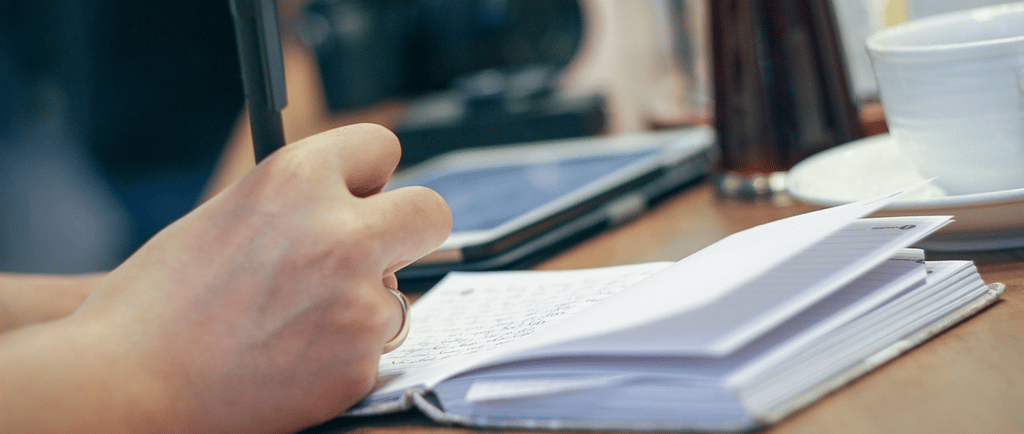 Person writing in a journal, with a cup placed nearby