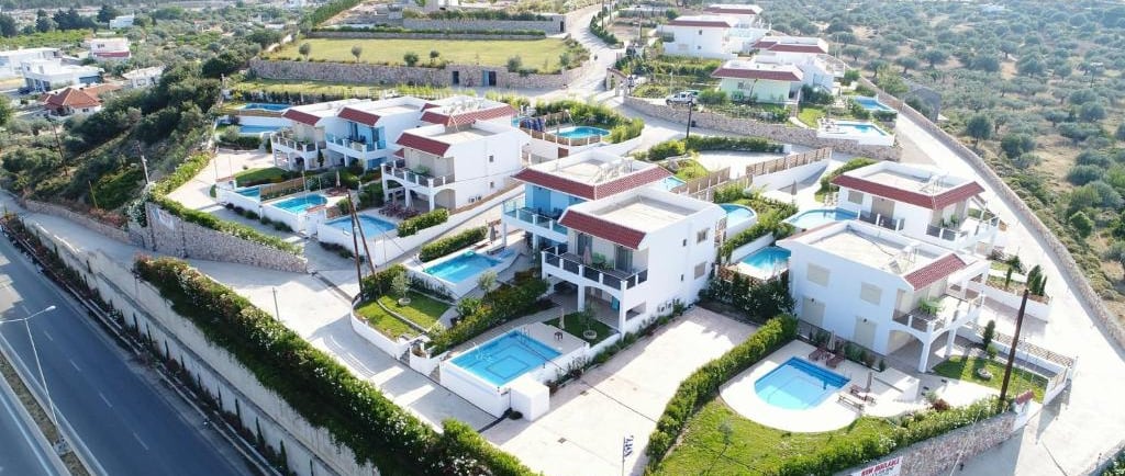 Aerial view of luxury vacation villas with private swimming pools in a hillside Greek resort.