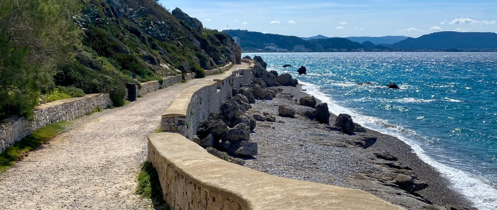 A scenic coastal walking path along a rocky beach with blue ocean water under a clear sky.