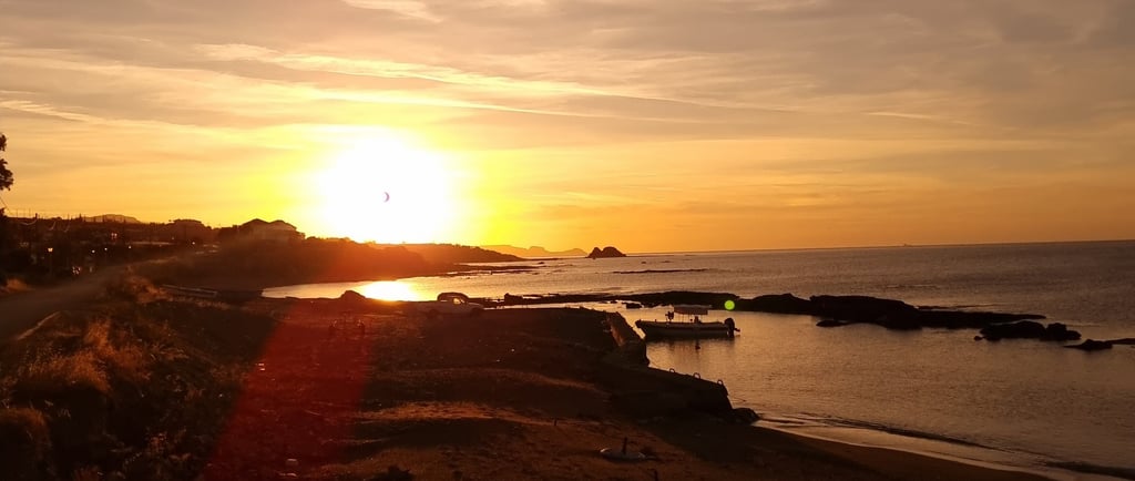 Golden sunset over a rocky coastline with a calm ocean beach and a small boat at dusk.