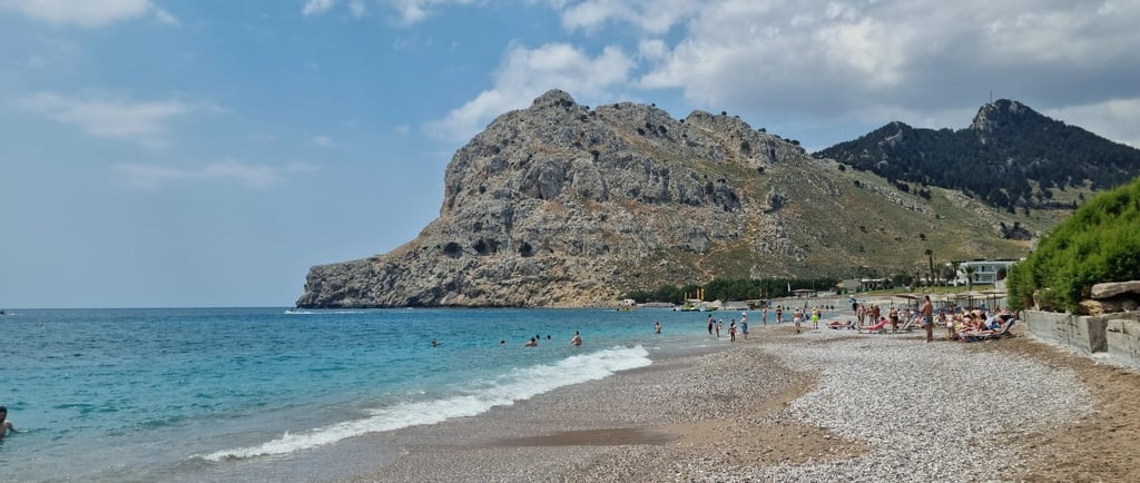 Sunny pebble beach in Rhodes with tourists swimming in turquoise water near a large rocky mountain.