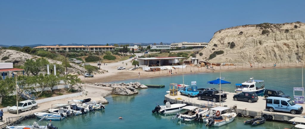 Scenic Mediterranean harbor with motorboats docked in turquoise water near a sandy beach and rocky cliffs.